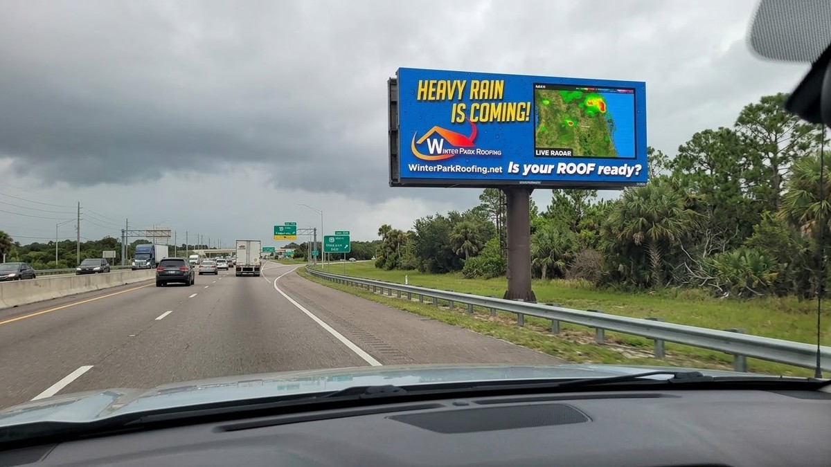 Highway digital billboard showing a live weather radar alert paired with a Winter Park Roofing ad